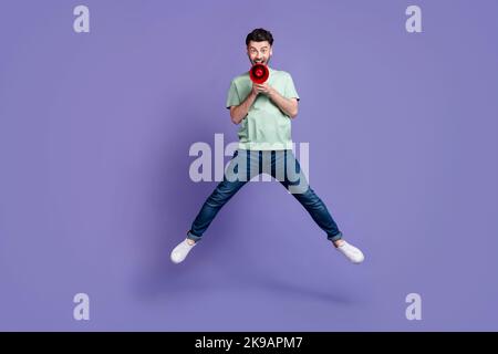 Photo pleine longueur d'un jeune homme gentil sautant haut tenue mégaphone criant habillé élégant vêtement gris isolé sur fond violet de couleur Banque D'Images