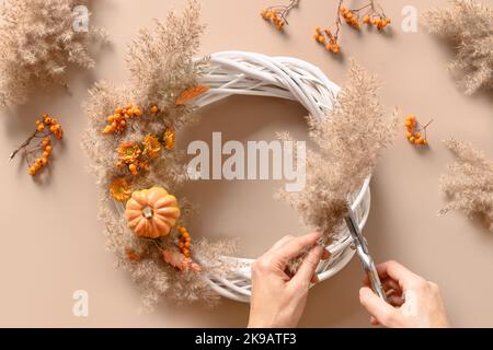 Femme en train de faire une couronne avec des fleurs orange et des matériaux naturels secs sur fond beige. Vue de dessus. Atelier pour la décoration artisanale de Thanksgiving. Banque D'Images