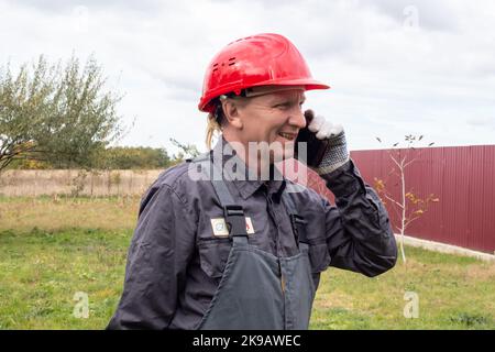 Un homme adulte travaillant en combinaison et un casque de sécurité parle au téléphone dans la rue et sourit. Banque D'Images