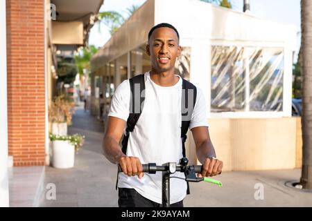 Joyeux homme afro-américain souriant dans des vêtements décontractés qui se déplace un scooter électrique dans la rue par une journée ensoleillée. Joyeux homme beau à conduire un scooter Banque D'Images