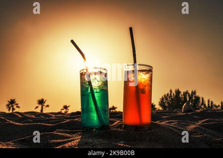 Boissons alcoolisées colorées avec une paille dans le sable avec des palmiers à l'arrière-plan avec le ciel de coucher du soleil Banque D'Images