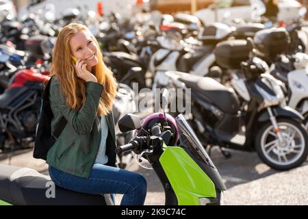 Femme aux cheveux rouges sur sa moto dans un parking parlant sur un smartphone Banque D'Images