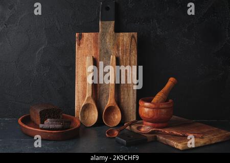Planches à découper en bois, mortier avec pilon, cuillères et pain sur fond noir Banque D'Images