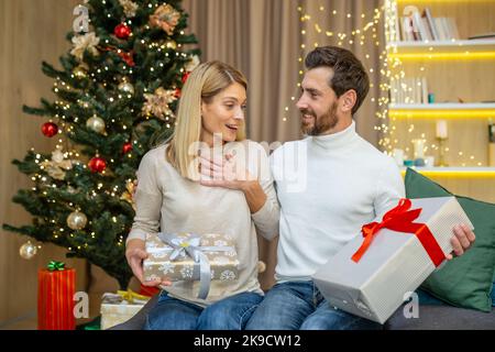 Couple de famille en amour homme et femme à Noël célébrant les vacances du nouvel an et échanger des cadeaux assis sur le canapé à la maison près de l'arbre de Noël dans la salle de séjour. Banque D'Images