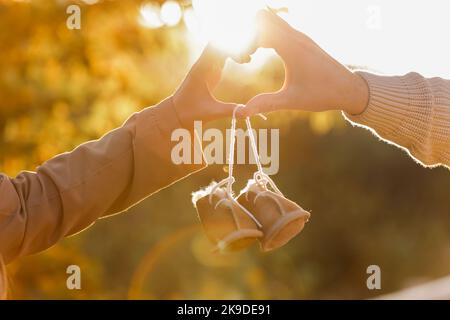 L'homme et la femme enceinte font des gestes cardiaques avec les mains et tiennent des chaussures chaudes pour bébé sur fond d'automne nature. Futurs parents attendant le bébé. Le concept de la Fête des mères et de la Journée des femmes Banque D'Images