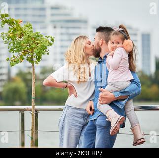 Homme et femme avec enfant à l'extérieur. Homme barbu tenant une petite fille et embrassant une femme blonde tout en passant du temps avec la famille dans le nouveau quartier urbain. Banque D'Images