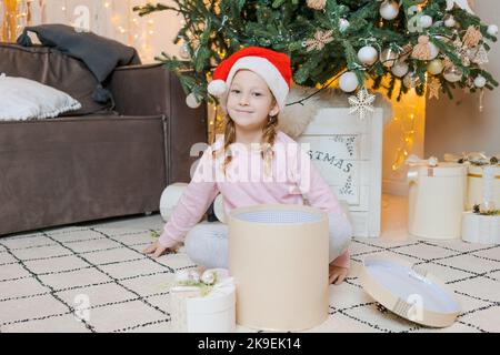 La fillette en pyjama est accompagnée d'un cadeau dans un chapeau du père noël dans une chambre décorée pour noël. Des vacances d'hiver fabuleuses. Concept de noël et de la bonne enfance de la nouvelle année Banque D'Images