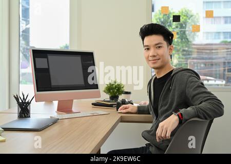 De beaux éditeurs de photos masculins assis dans un lieu de travail créatif devant un ordinateur personnel et souriant à l'appareil photo Banque D'Images