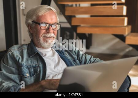 Un homme âgé avec des lunettes aux cheveux gris et une barbe est assis à la maison sur le canapé ou parcourt les réseaux sociaux sur un ordinateur portable Banque D'Images