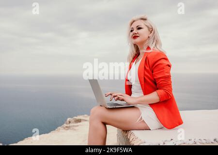 Digital nomade, une femme d'affaires dans un blazer rouge avec un ordinateur portable se trouve sur les rochers au bord de la mer pendant le coucher du soleil, effectue une transaction d'affaires en ligne à partir d'un Banque D'Images