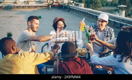 De jeunes amis gaies discutent et boivent de la limonade sur le toit lors d'une chaude journée ensoleillée avec des en-cas et des boissons sur la table. Concept été, jeunesse et fête. Banque D'Images