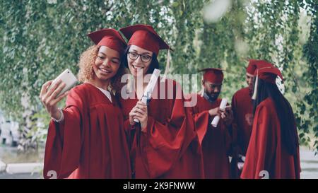 Les jolies femmes diplômées prennent le selfie avec le défilement du diplôme en utilisant le smartphone, les jeunes femmes posent avec d'autres étudiants dans les robes mobiles et parler en arrière-plan. Banque D'Images