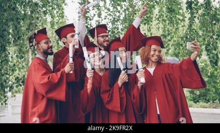 Les jeunes diplômés enthousiastes prennent le selfie avec leur smartphone, les jeunes font des diplômes, posent, sourient et rient. Éducation, jeunesse et réussite. Banque D'Images