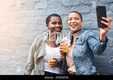 Discutez en direct n'importe où, n'importe quand. Deux jeunes femmes se tenant à côté d'un bâtiment souriant et prenant des selfies tout en tenant leurs boissons fraîches. Banque D'Images
