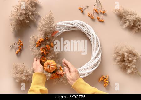 Femme en train de faire une couronne avec des fleurs orange et des matériaux naturels secs sur fond beige. Vue de dessus. Atelier pour la décoration artisanale de Thanksgiving. Banque D'Images