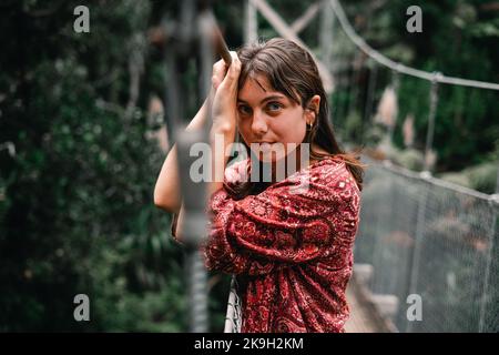 jeune femme caucasienne avec des yeux bleus et un nez étroit regardant la caméra et s'appuyant sur la corde métallique du long et étroit pont qui traverse Banque D'Images