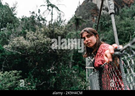 jeune femme caucasienne aux yeux bleus, nez étroit avec boucles d'oreilles rondes et vêtements larges, penchée sur la corde métallique du pont qui traverse Banque D'Images