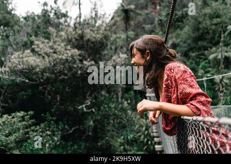 caucasienne fille avec des cheveux bruns salissants souriez détendu s'appuyant sur la main courante en métal du pont de suspension qui traverse la rivière de la forêt en donut Banque D'Images