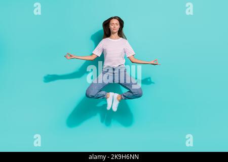 Photo pleine grandeur d'une femme calme et détendue avec des cheveux bruns porter un t-shirt rayé lévitant dans le yoga pose isolée sur fond de couleur sarcelle Banque D'Images