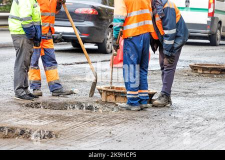 Les travailleurs de la route réparent les vieux trous d'égout sur la chaussée contre le fond des voitures qui passent. Banque D'Images