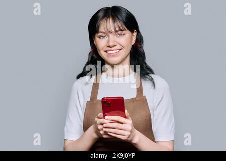 Souriant jeune femme dans un tablier à l'aide d'un smartphone, sur fond gris Banque D'Images