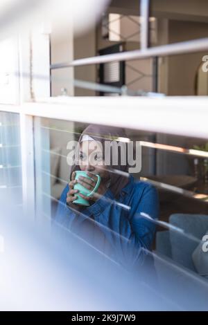 Femme biraciale réfléchie portant le hijab, regardant à travers la fenêtre et buvant du café Banque D'Images