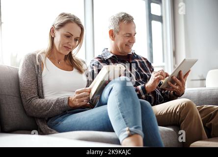 Le dimanche devrait être la manière. Une femme mûre lisant un livre tandis que son mari utilise une tablette numérique sur le canapé à la maison. Banque D'Images