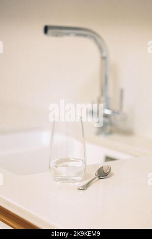 Verre d'eau et cuillère à café sur table dans la cuisine avec robinet, gros plan. Photo de haute qualité Banque D'Images