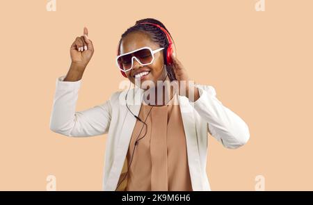 Joyeuse femme afro-américaine aime la musique dans les écouteurs et la danse sur fond beige. Banque D'Images