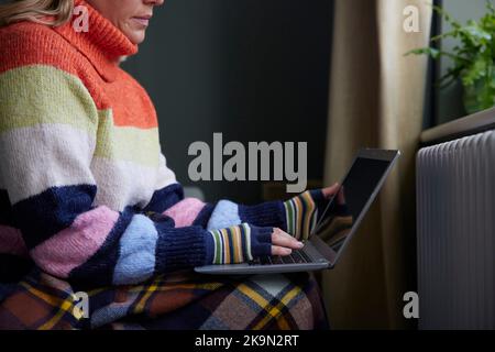 Femme en gants avec ordinateur portable essayant de rester au chaud par le radiateur pendant la crise énergétique du coût de la vie Banque D'Images