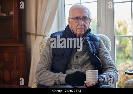 Portrait de l'homme senior portant des vêtements supplémentaires avec boisson chaude essayant de rester au chaud à la maison en crise d'énergie Banque D'Images