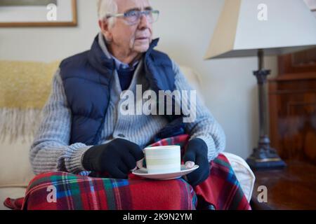 Homme senior portant des vêtements supplémentaires avec boisson chaude essayant de rester au chaud à la maison en crise d'énergie Banque D'Images