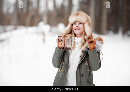 Belle femme souriante admirant la nature hivernale dans la forêt, portant veste kaki en fourrure, rabats d'oreilles chapeau et gants en cuir, portrait confortable sur fond de neige Banque D'Images