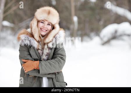 Portrait de la belle fille heureuse debout dans la nature hiver neige forêt. Femme souriante portant des vêtements chauds, veste de parka d'hiver, chapeau de fourrure, gants en cuir Banque D'Images