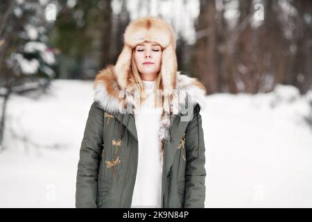 Femme aux yeux fermés dans la forêt d'hiver. Jeune femme portrait léger pendant la relaxation et la méditation, porter un chapeau de fourrure et une veste de parka, pratiquer le yoga ou Banque D'Images