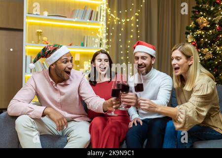 Jeunes gens de groupe joyeux, amis fêtent le compte à rebours du nouvel an à la maison clin d'œil des verres à vin tout en étant assis sur le canapé à la maison. Ils se souhaitent un joyeux Noël heureux et un câlin ensemble d'avoir du plaisir Banque D'Images