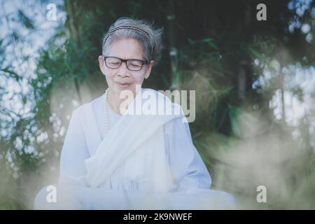 Calme de la femme asiatique âgée en robe blanche avec des cheveux blancs assis pour respirer la méditation sur fond vert nature dans le parc Banque D'Images