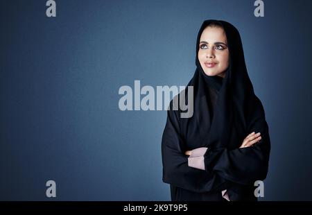 Rêver d'un avenir réussi. Une jeune femme d'affaires attirante debout avec ses bras repliée sur un fond sombre. Banque D'Images