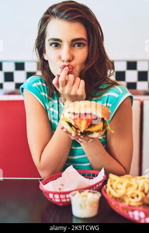 La perfection. Portrait court d'une jeune femme attrayante qui apprécie un hamburger dans un restaurant rétro. Banque D'Images