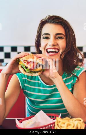 Ils font les meilleurs hamburgers. Portrait court d'une jeune femme attrayante qui apprécie un hamburger dans un restaurant rétro. Banque D'Images