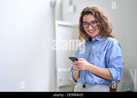 Jeune femme d'affaires heureuse utilisant un téléphone portable debout dans le bureau utilisant le mobile. Banque D'Images