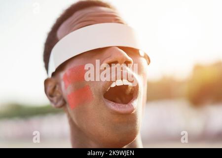 Fan de sport rouge africain regardant un match de football en dehors du stade avec des lunettes futuristes de réalité augmentée - Focus on face Banque D'Images
