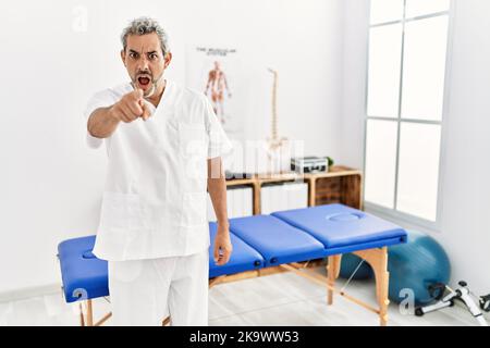 Thérapeute hispanique d'âge moyen, homme travaillant à la clinique de rétablissement de la douleur, pointant mécontent et frustré à la caméra, en colère et furieux avec vous Banque D'Images