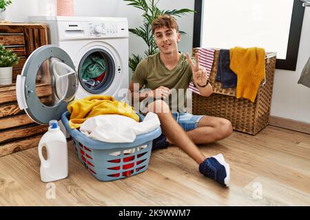 Jeune homme caucasien mettant le linge sale dans la machine à laver souriant regardant la caméra montrant les doigts faisant signe de victoire. Numéro deux. Banque D'Images