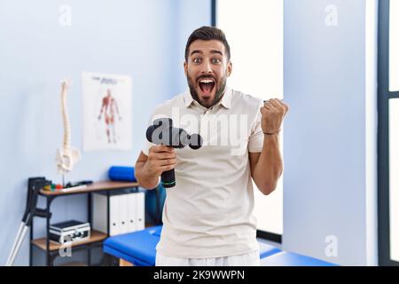 Beau homme hispanique tenant un pistolet de massage thérapeutique au centre de physiothérapie criant fier, célébrant la victoire et le succès très excité avec ar élevé Banque D'Images