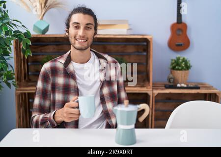 Un jeune hispanique boit un café de la cafetière française, en train de se tenir debout et souriant avec un sourire confiant montrant des dents Banque D'Images
