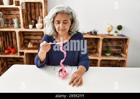 Femme d'âge moyen à cheveux gris mangeant un mètre-ruban assis sur la table à la maison. Banque D'Images