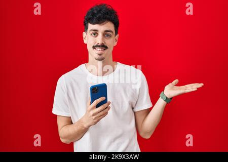 Homme hispanique utilisant un smartphone sur fond rouge pointant de côté avec les mains ouvertes palmiers montrant l'espace de copie, présentant la publicité souriant ha excité Banque D'Images