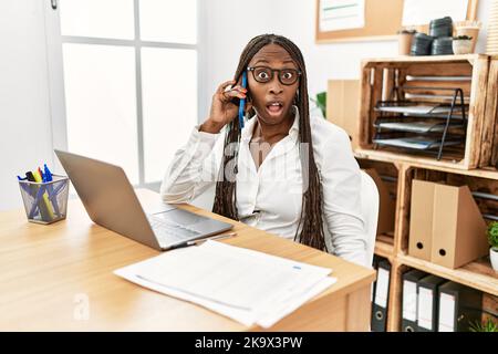 Femme noire avec des tresses travaillant au bureau parlant au téléphone dans le visage de choc, regardant sceptique et sarcastique, surpris par la bouche ouverte Banque D'Images