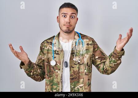 Jeune médecin hispanique portant l'uniforme de l'armée de camouflage criant et criant à voix haute à côté de la main sur la bouche. Concept de communication. Banque D'Images